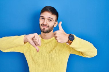 Latin kökenli bir adam mavi arka planda dikiliyor. Başparmak kaldırıyor, anlaşmazlık ve anlaşma ifadesi takınıyor. çılgın çatışma 