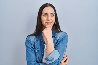 Mavi arka planda duran İspanyol kadın kameraya kendinden emin bir şekilde bakıyor kollarını kavuşturmuş ve çenesini kaldırmış gülümsüyor. Olumlu düşünmek. 