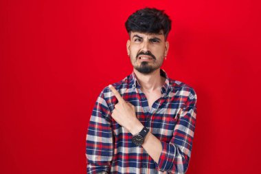 Kırmızı arka planda duran sakallı genç İspanyol adam endişeli ve endişeli bir ifade takınıyor. 