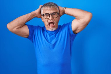 Mavi arka planda gri saçlı İspanyol bir adam. Deli ve elleri başında korkmuş, ağzı açık şoktan korkmuş ve şaşırmış. 