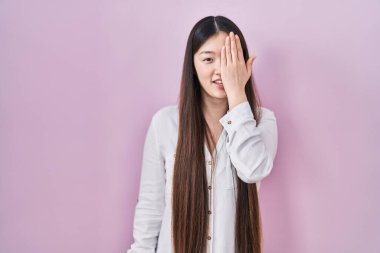 Chinese young woman standing over pink background covering one eye with hand, confident smile on face and surprise emotion. 