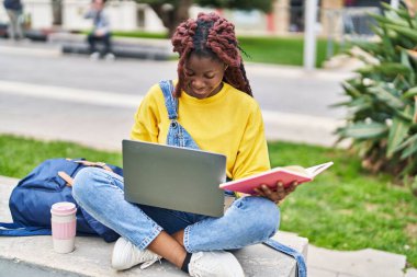 African american woman student using laptop reading book at campus park