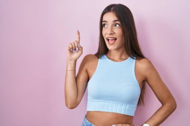 Young brunette woman standing over pink background pointing finger up with successful idea. exited and happy. number one. 