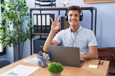 Ofiste çalışan Latin asıllı genç adam kulaklık takıyor ve üç numaralı parmaklarıyla işaret ederken kendinden emin ve mutlu bir şekilde gülümsüyor.. 