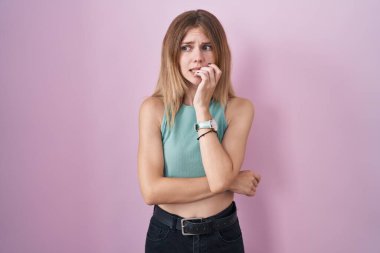 Blonde caucasian woman standing over pink background looking stressed and nervous with hands on mouth biting nails. anxiety problem. 