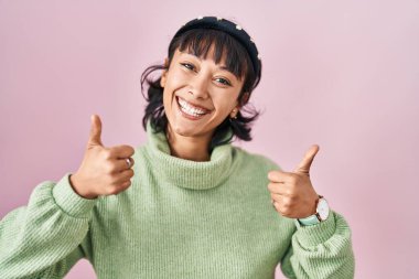 Young beautiful woman standing over pink background success sign doing positive gesture with hand, thumbs up smiling and happy. cheerful expression and winner gesture. 