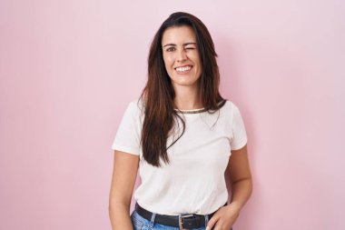 Genç esmer kadın pembe arka planda duruyor. Seksi bir ifadeyle kameraya bakıyor. Neşeli ve mutlu bir yüzü var.. 