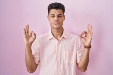 Pembe arka planda duran genç İspanyol adam rahatlamış ve gözleri kapalı gülümsüyor parmaklarıyla meditasyon hareketi yapıyor. yoga konsepti. 