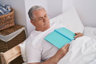 Orta yaşlı, kır saçlı, yatakta uzanmış, elinde kitap tutan bir adam.