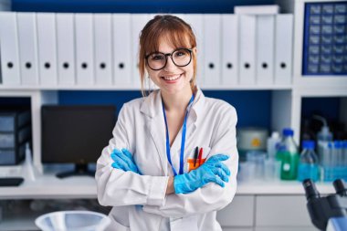 Genç bilim kadını gülümsüyor. Kollarını kavuşturup laboratuvarda duruyor.