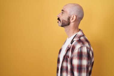 Hispanic man with beard standing over yellow background looking to side, relax profile pose with natural face with confident smile. 