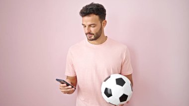 Genç İspanyol adam, topu pembe arka planda tutarken akıllı telefon kullanıyor.