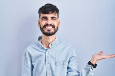 Mavi arka planda sakallı, neşeli bir şekilde gülümseyen ve avuç içi kameraya bakan genç İspanyol bir adam.. 