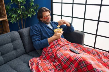 Orta yaşlı bir adam göz bandı takıyor ve evde patates cipsi yiyor.