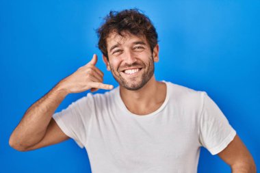 İspanyol genç adam mavi arka planda durup gülümsüyor telefonla el ve parmaklarıyla telefon hareketi yapıyor telefonda konuşuyor gibi. iletişim kavramları. 