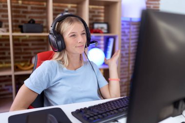 Video oyunları oynayan, kulaklık takan, neşeli bir şekilde gülümseyen ve avuç içi kameraya bakan beyaz bir kadın.. 