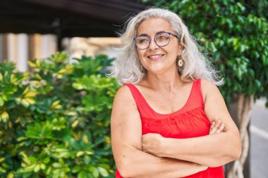 Orta yaşlı, kır saçlı, parkta kollarını kavuşturmuş bir kadın.