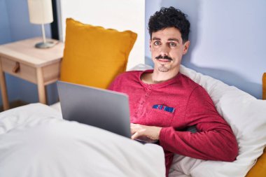 Yatak odasında dizüstü bilgisayar kullanan genç beyaz bir adam.