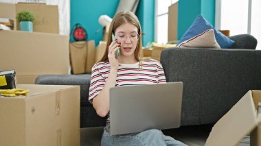 Genç sarışın kadın dizüstü bilgisayar kullanıyor. Yeni evinde akıllı telefondan konuşuyor.