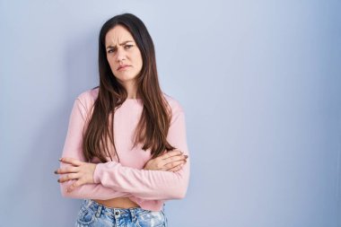 Mavi arka planda duran esmer kadın şüpheci ve gergin, yüzünde çapraz kollu tasvip etmeyen ifade. negatif kişi. 