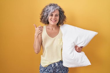 Orta yaşlı, gri saçlı, pijama giyen, yastığa sarılan mutlu baş parmaklı el hareketi yapan bir kadın. Kameraya bakıp başarıyı gösteren ifade. 