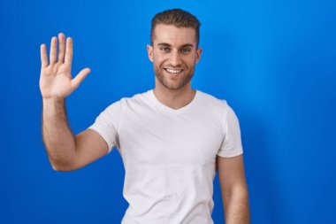 Mavi arka planda duran genç beyaz adam merhaba diyor mutlu ve gülümseyen, dostça bir hoş geldin jesti. 