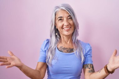 Pembe arka planda duran orta yaşlı bir kadın. Kollarını açıp gülümsüyor. Dostça karşılama, olumlu ve kendinden emin selamlar. 