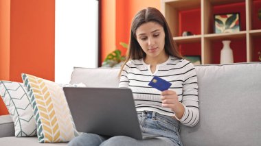 Laptop ve kredi kartıyla alışveriş yapan genç, güzel İspanyol kadın evdeki kanepede oturuyor.