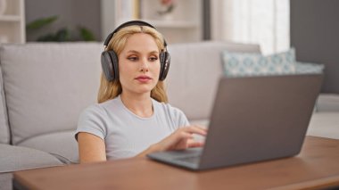 Dizüstü bilgisayar ve kulaklık kullanan genç sarışın kadın evde oturuyor.
