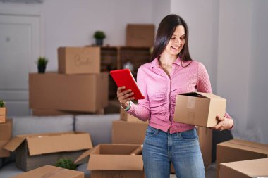 Young beautiful hispanic woman using touchpad holding package at new home