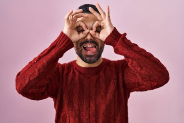 Sakallı, pembe arka planda rahat süveter giyen genç İspanyol bir adam dürbün gibi dil çıkartıyor, gözler parmaklarından bakıyor. çılgın ifade. 