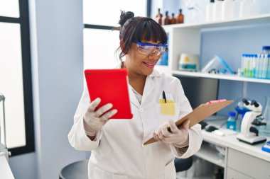 Young beautiful latin woman scientist using touchpad reading document at laboratory