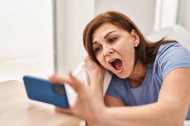 Orta yaşlı bilim kadını gülümsüyor. Kollarını kavuşturup yatak odasında oturuyor.