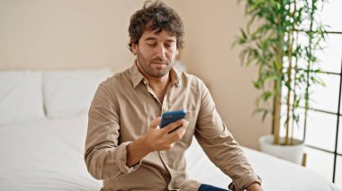 Genç İspanyol adam yatakta akıllı telefon kullanıyor.