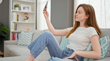 Sarışın bir kadın akıllı telefonuyla fotoğraf çekiyor. Evdeki kanepede oturuyor.