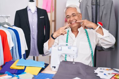 Orta yaşlı bir adam, gri kuaförüyle dikiş makinesi kullanıyor neşeli bir şekilde gülümsüyor ve parmaklarını ve ağzını işaret ediyor. diş sağlığı kavramı. 