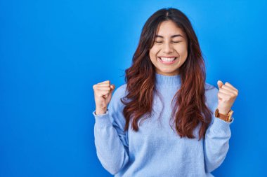 Mavi arka planda duran Latin kökenli genç bir kadın başarı için heyecanlı kolları havada ve gözleri kapalı zaferi kutlarken gülümsüyor. Kazanan konsept. 