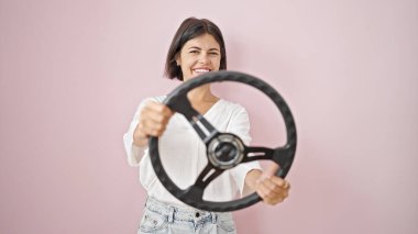 Genç, güzel, İspanyol bir kadın, kendine güvenen, direksiyon simidini, izole edilmiş pembe arka planda kullanan.