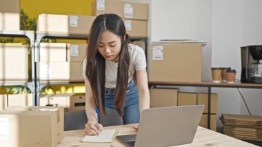 Genç Çinli kadın ekommerce işçisi ofiste dizüstü bilgisayar kullanarak notlar yazıyor.
