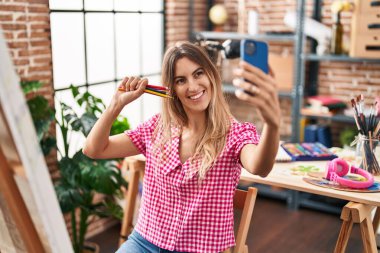 Genç kadın sanatçı, resim stüdyosunda fırça tutarak akıllı telefondan selfie çekiyor.