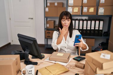 Küçük bir iş yerinde çalışan esmer bir kadın akıllı telefon kullanarak ağzını eliyle kapatıyor, şok geçiriyor ve hata yapmaktan korkuyor. şaşırmış ifade 