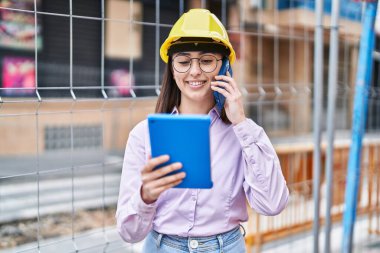 Young hispanic woman architect talking on the smartphone using touchpad at street