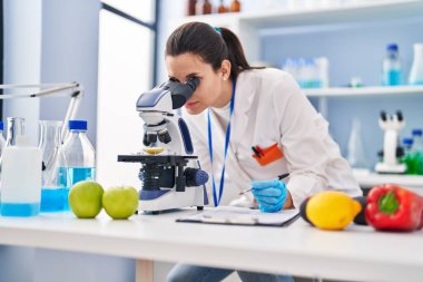 Young beautiful hispanic woman scientist using microscope writing on document at laboratory