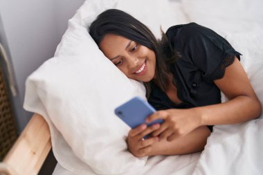 Genç İspanyol kadın yatakta akıllı telefon kullanıyor.