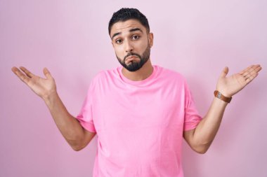 Pembe arka planda duran Latin kökenli genç adam elleri ve kolları havada, kafası karışmış bir ifade takınıyor. şüphe kavramı. 