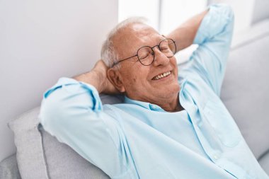 Yaşlı, kır saçlı, elleri başının üstünde rahat bir şekilde evdeki kanepede oturuyor.