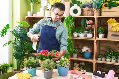 Young hispanic man florist using diffuser watering plant at flower shop