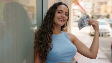 Young beautiful hispanic woman smiling confident using dollars as a handfan at street