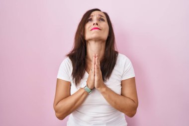 Orta yaşlı esmer kadın pembe arka planda duruyor. Elleriyle yalvarıyor ve dua ediyor. Yüzünde umut ifadesi var. Yalvarıyor. 