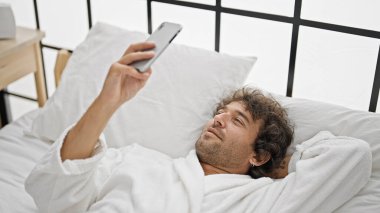 Genç İspanyol adam bornoz giyiyor. Yatak odasında akıllı telefon kullanıyor.
